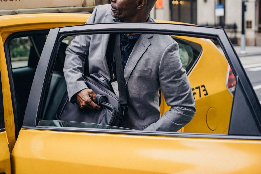 Crop African American male in formal suit sitting in yellow taxi car parked on city street